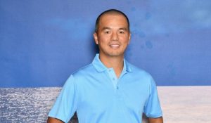mike chen in a blue shirt stands against a solid blue background, creating a monochromatic look