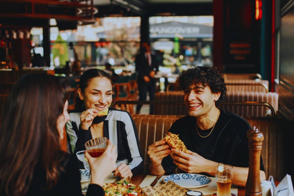 Three friends meet at a restaurant, eating pizza and toasting with wine, enjoying their time together.