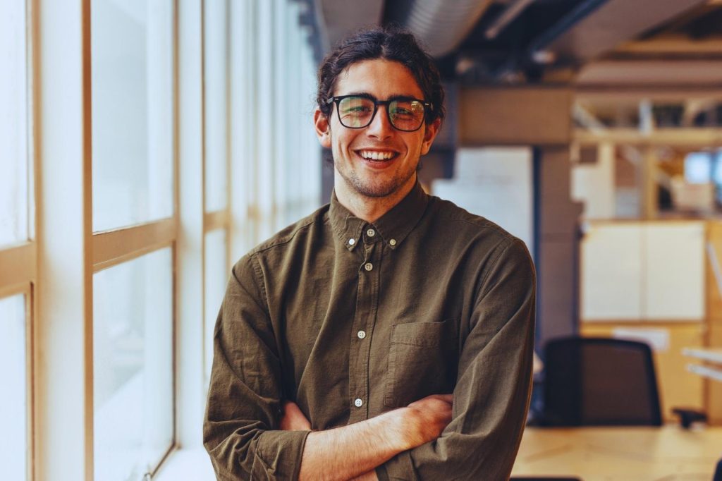 Man with glasses, standing by a window, attentively observing the outside landscape.
