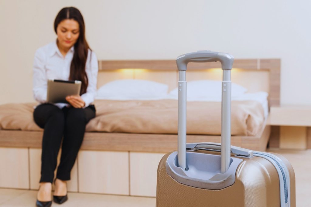 Mujer sentada en una cama con una maleta a su lado, mirando hacia el frente con una expresión pensativa.