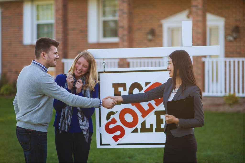Two people shake hands in front of a sign that says "sold".