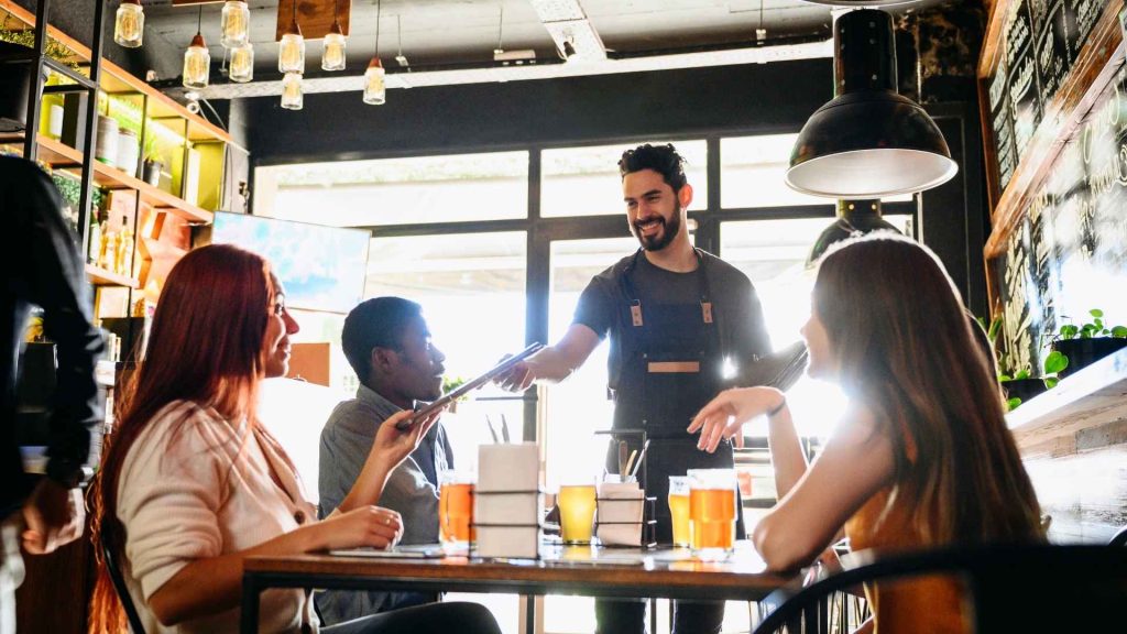 Camarero sonriente entregando la carta a un grupo de amigos en un restaurante moderno con iluminación cálida