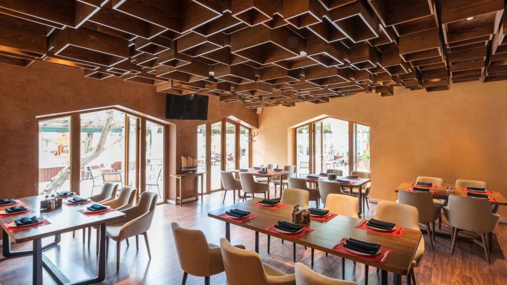 Interior de un restaurante moderno con un techo artístico de cubos de madera, mesas preparadas para el servicio y grandes ventanales con luz natural.