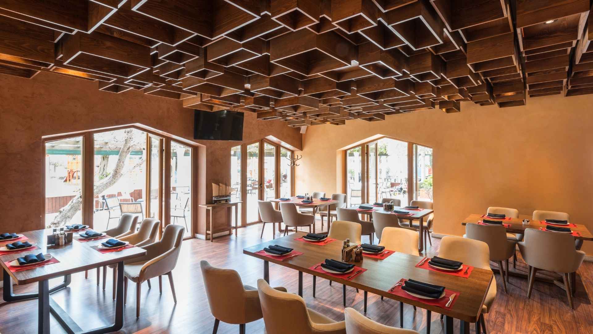 Interior de un restaurante moderno con un techo artístico de cubos de madera, mesas preparadas para el servicio y grandes ventanales con luz natural.