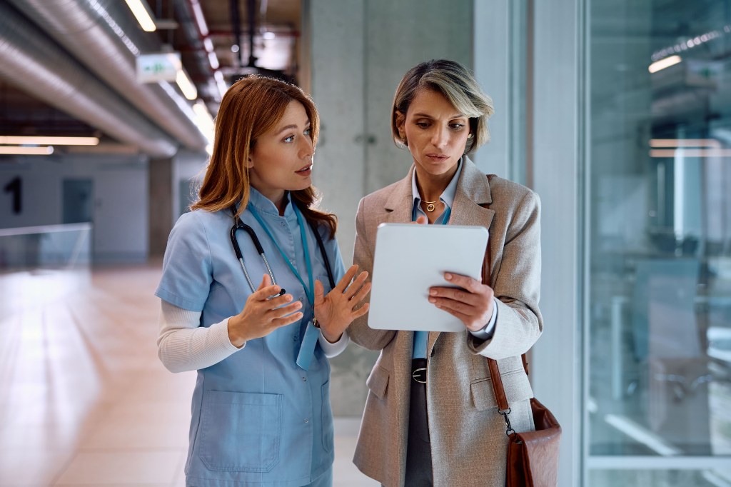 Una enfermera con uniforme azul y estetoscopio gesticula mientras explica información en una tableta digital a una administradora de hospital