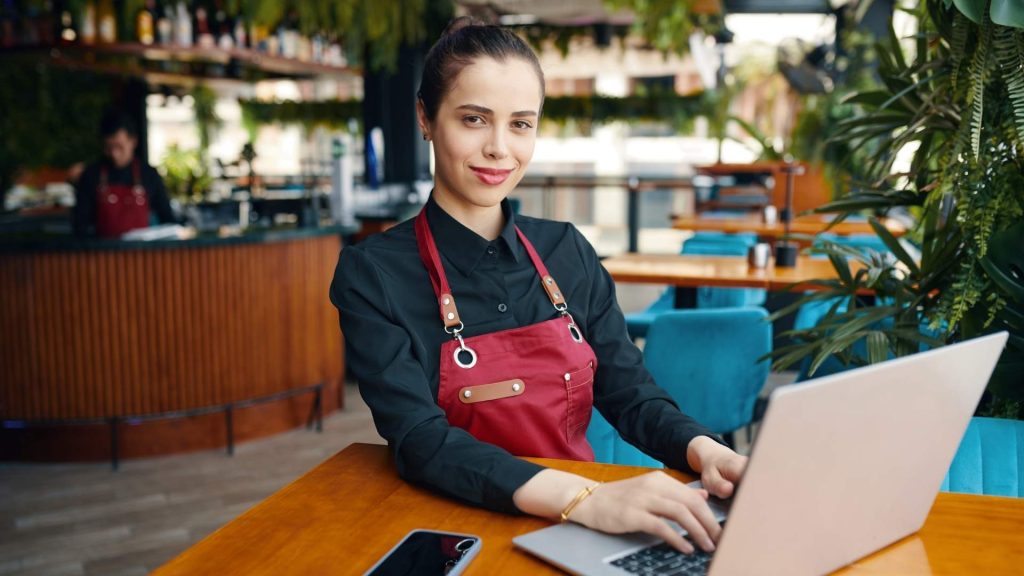 Mujer joven con delantal de camarera trabajando con un ordenador portátil en la mesa de un restaurante moderno.
