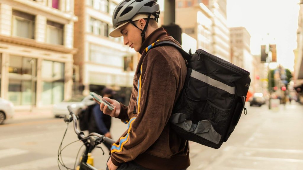 Repartidor de delivery con mochila térmica y casco revisando su teléfono móvil en una calle transitada.