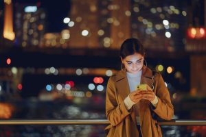 Mujer usando su smartphone de noche en la ciudad, representando búsquedas de último minuto para reservas de hotel.