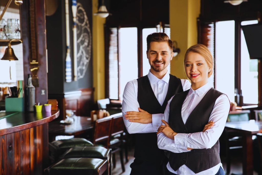 Pareja de personal de servicio uniformado con chaleco y camisa blanca.