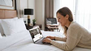Mujer sonriente acostada en una cama de hotel usando una computadora portátil que muestra el portal de reservas reserva directa.