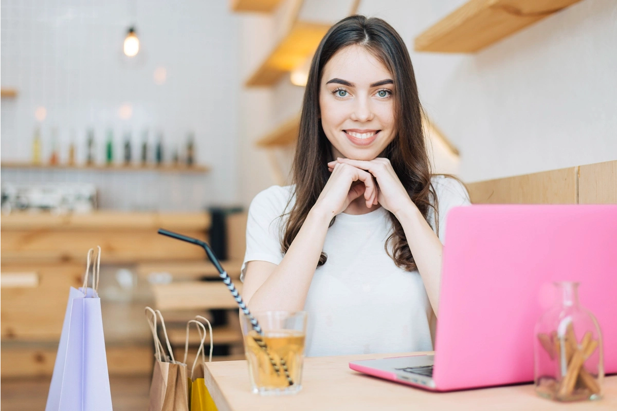 Compradora sonriente con laptop y bolsas de compras en café.